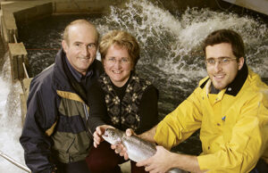 Trois personnes regroupées près d'un bassin d'élevage de truites, l'une tenant une truite fraîchement pêchée - pisciculture des Bobines au Québec