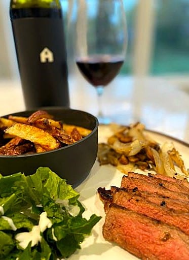 Bavette tranchée dans une assiette, accompagnée d'une salade de pommes de terre style grecque et d'un verre de vin rouge avec sa bouteille — un repas gourmet Pour Le Chef