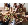 Top-down view of a group of people seated around a wooden table, sharing a meal with bowls and plates of salad and pasta in the center.