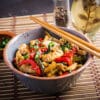 Bowl of chicken stir-fry with red peppers and greens, sesame seeds, and chopped scallions; chopsticks rest on the bowl's edge.
