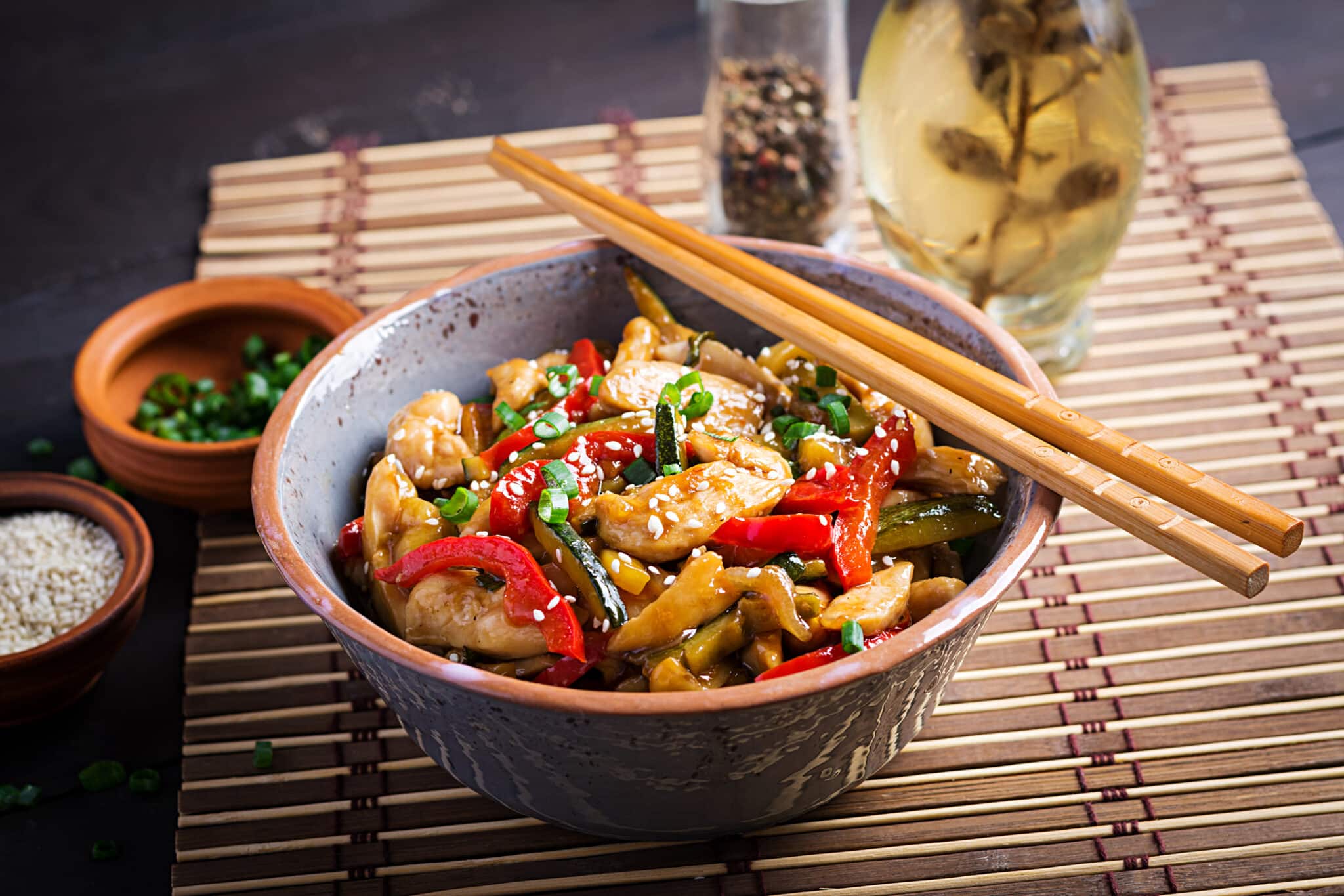 Bowl of chicken stir-fry with red peppers and greens, sesame seeds, and chopped scallions; chopsticks rest on the bowl's edge.