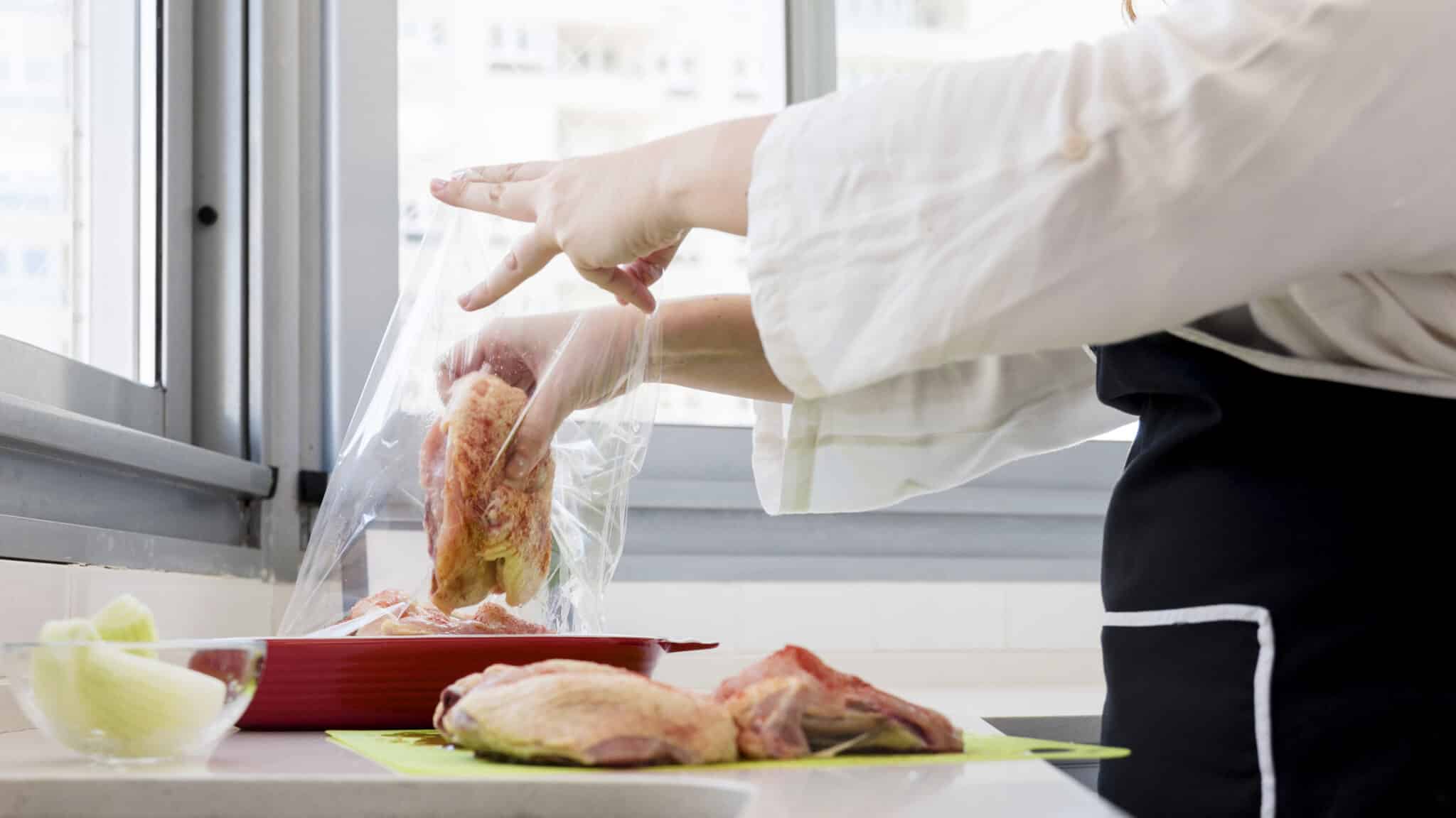 Person in a white coat packs baguette into a clear plastic bag on a kitchen counter near a window.