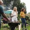 People enjoy a backyard barbecue: a man in a green shirt grills meat while a woman in a white star-printed top holds a plate among other guests chatting nearby by a grill and blue umbrella in a garden.