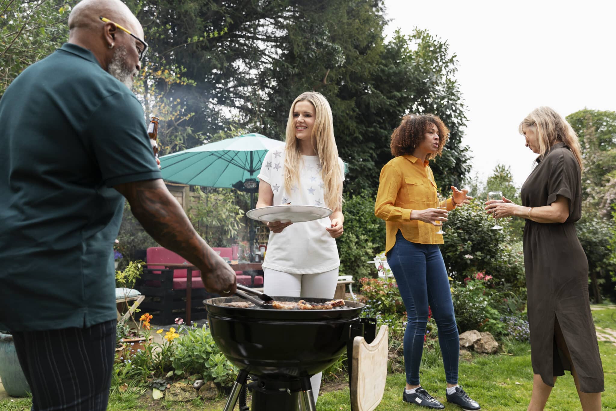 People enjoy a backyard barbecue: a man in a green shirt grills meat while a woman in a white star-printed top holds a plate among other guests chatting nearby by a grill and blue umbrella in a garden.