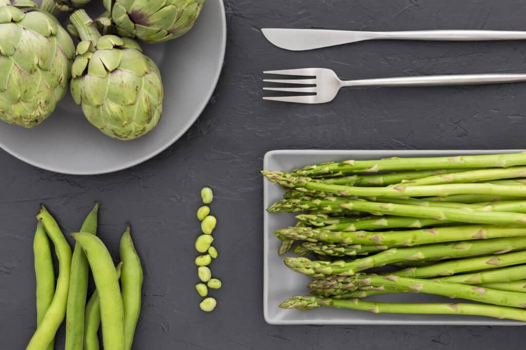 Fresh green asparagus spears laid out on a gray tray beside a plate of artichokes on a dark textured surface, with a knife and fork nearby.