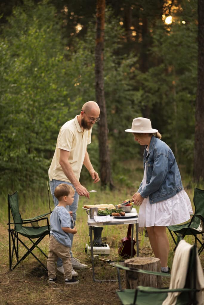 A family picnic in the woods: man, woman, and young boy cooking and serving food at a small outdoor table surrounded by camping chairs.