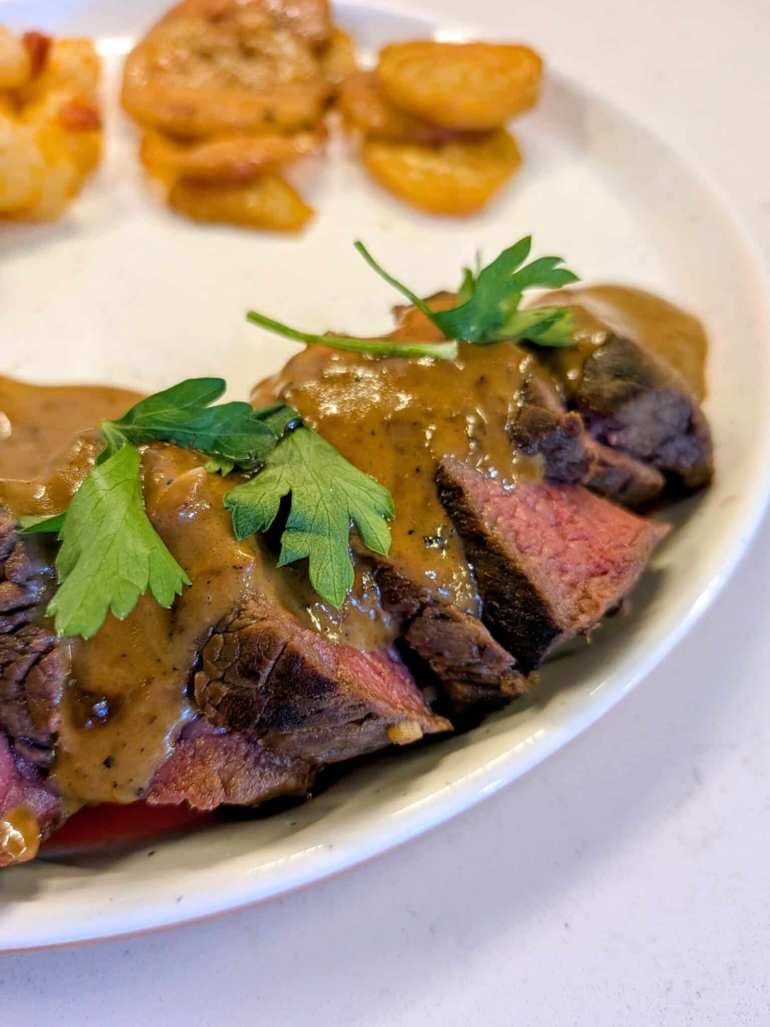 Sliced steak with brown sauce and fresh parsley garnish on a plate, with roasted potatoes in the background