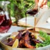 Plate of grilled meat and lemon wedges with fresh greens on a ceramic platter, outdoors on a sunny table.