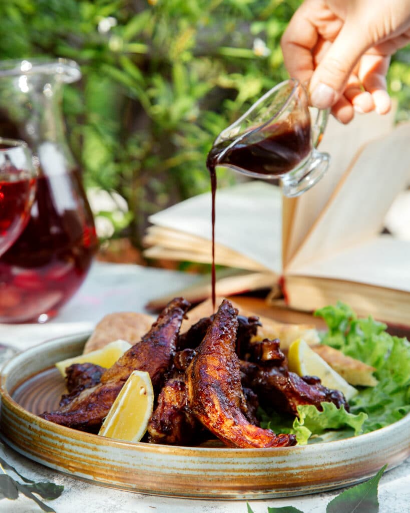 Plate of grilled meat and lemon wedges with fresh greens on a ceramic platter, outdoors on a sunny table.