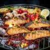 Salmon fillets grilling on a barbecue with lemon wedges, herbs, and vegetables over hot coals.