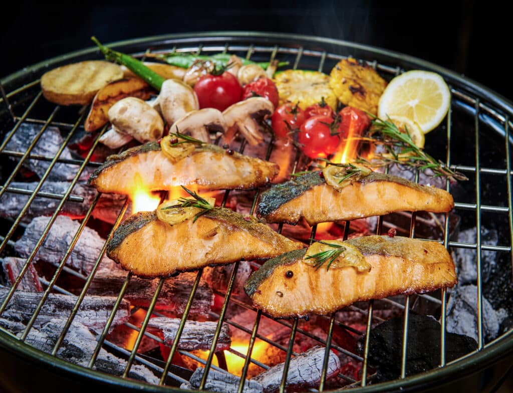 Salmon fillets grilling on a barbecue with lemon wedges, herbs, and vegetables over hot coals.