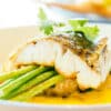 Seared fish fillet atop green asparagus on a plate with a yellow sauce and fresh parsley garnish