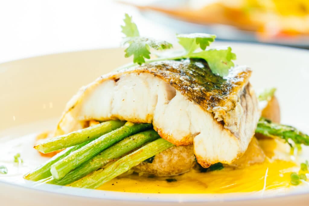 Seared fish fillet atop green asparagus on a plate with a yellow sauce and fresh parsley garnish