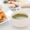 Plate of crispy fried chicken with a bowl of green soup and a white mug of coffee on a bright table surface.