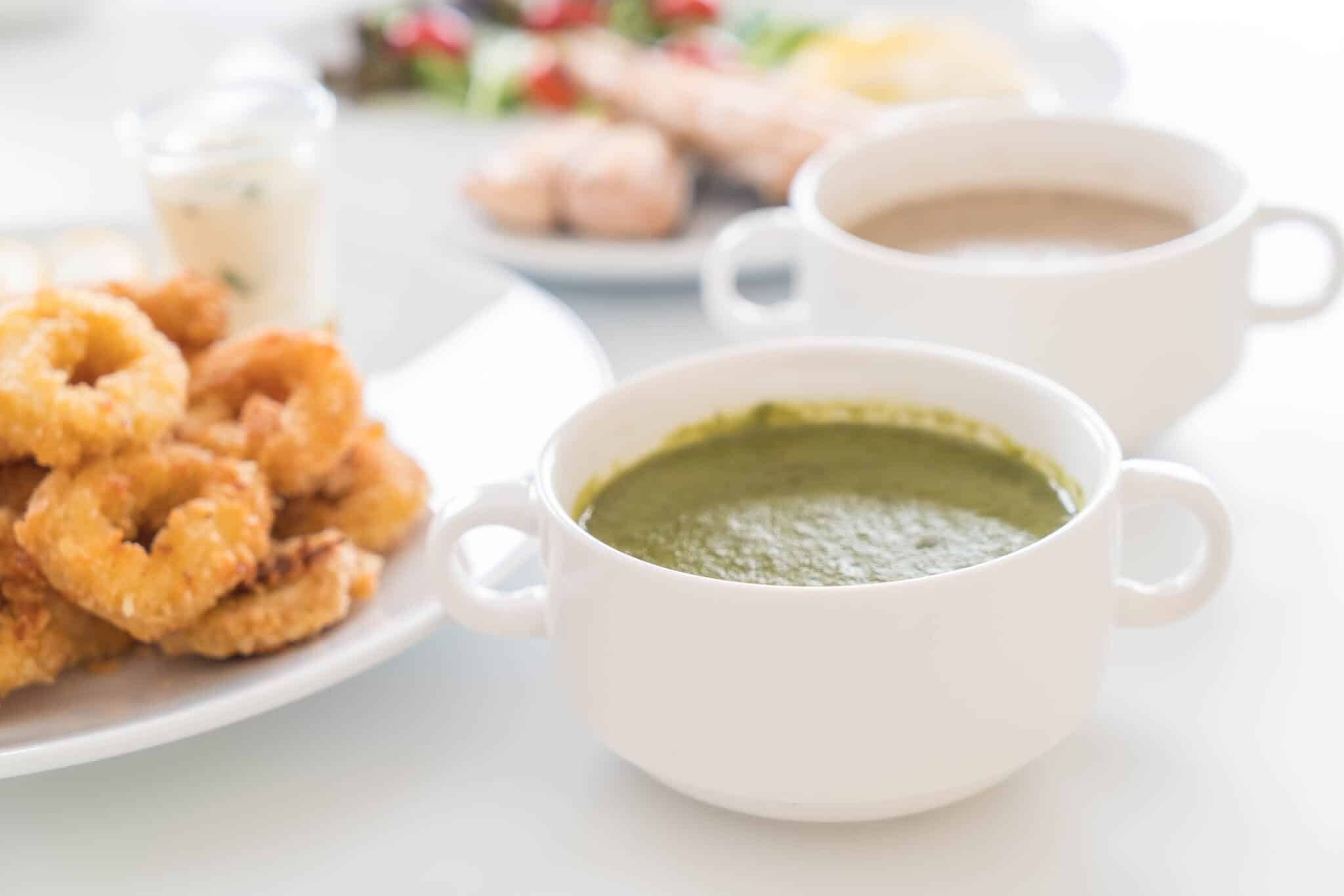 Plate of crispy fried chicken with a bowl of green soup and a white mug of coffee on a bright table surface.