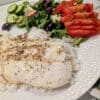 Plated white fish with peppercorn crust on rice, side salad with cucumber, olives, greens, and sliced tomatoes on a white plate.