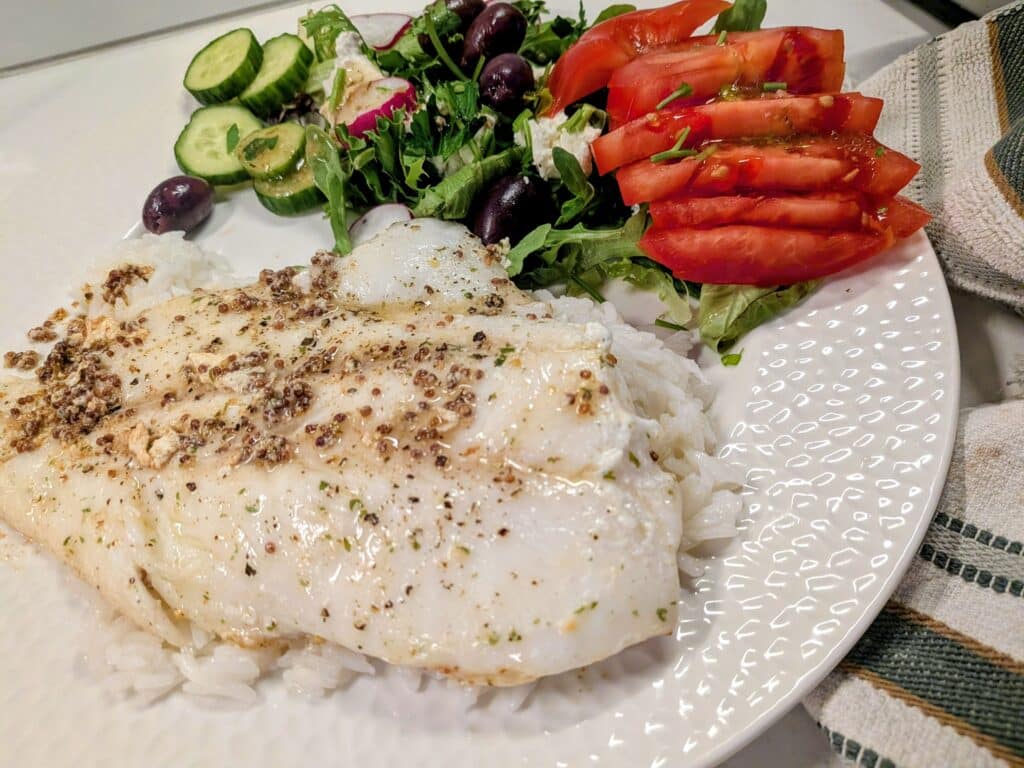 Plated white fish with peppercorn crust on rice, side salad with cucumber, olives, greens, and sliced tomatoes on a white plate.