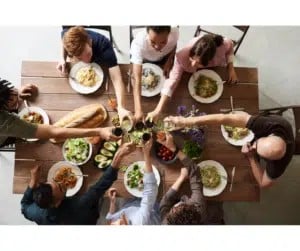 Top-down view of a diverse group of people seated around a wooden table, sharing a meal with bowls and plates of salad and pasta in the center.