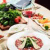 Plate of beef carpaccio topped with arugula and parmesan shavings on a dinner table, with a glass of red wine in the foreground.