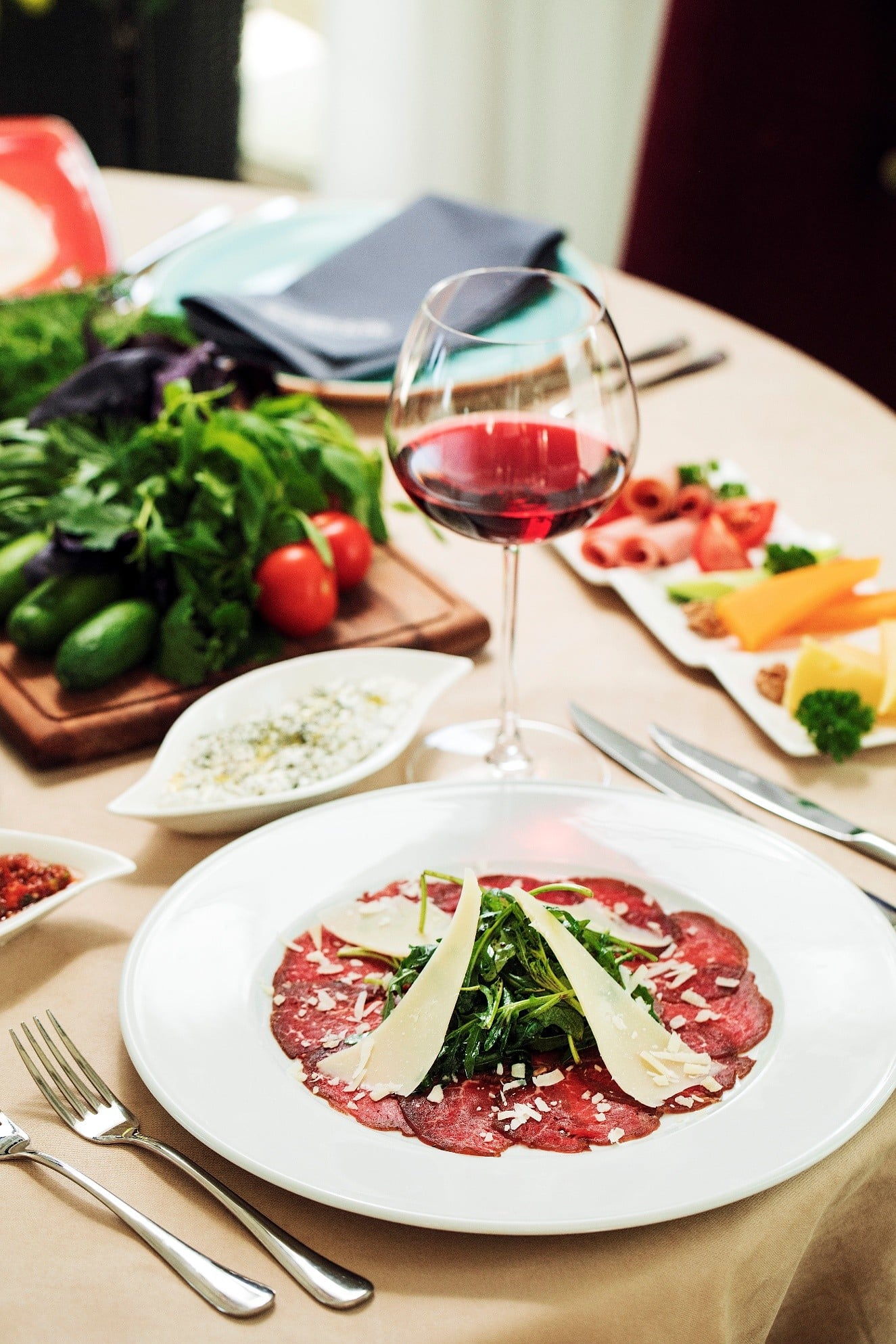Plate of beef carpaccio topped with arugula and parmesan shavings on a dinner table, with a glass of red wine in the foreground.