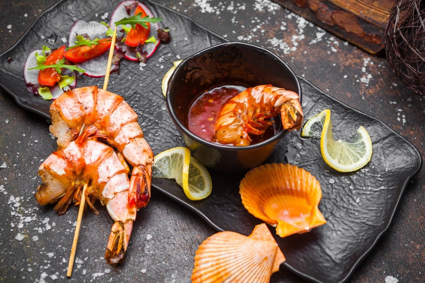 Platter of grilled shrimp with a dipping sauce in a black cup, lemon wedges, and seashells on a dark slate plate with garnish slices of radish and greens.