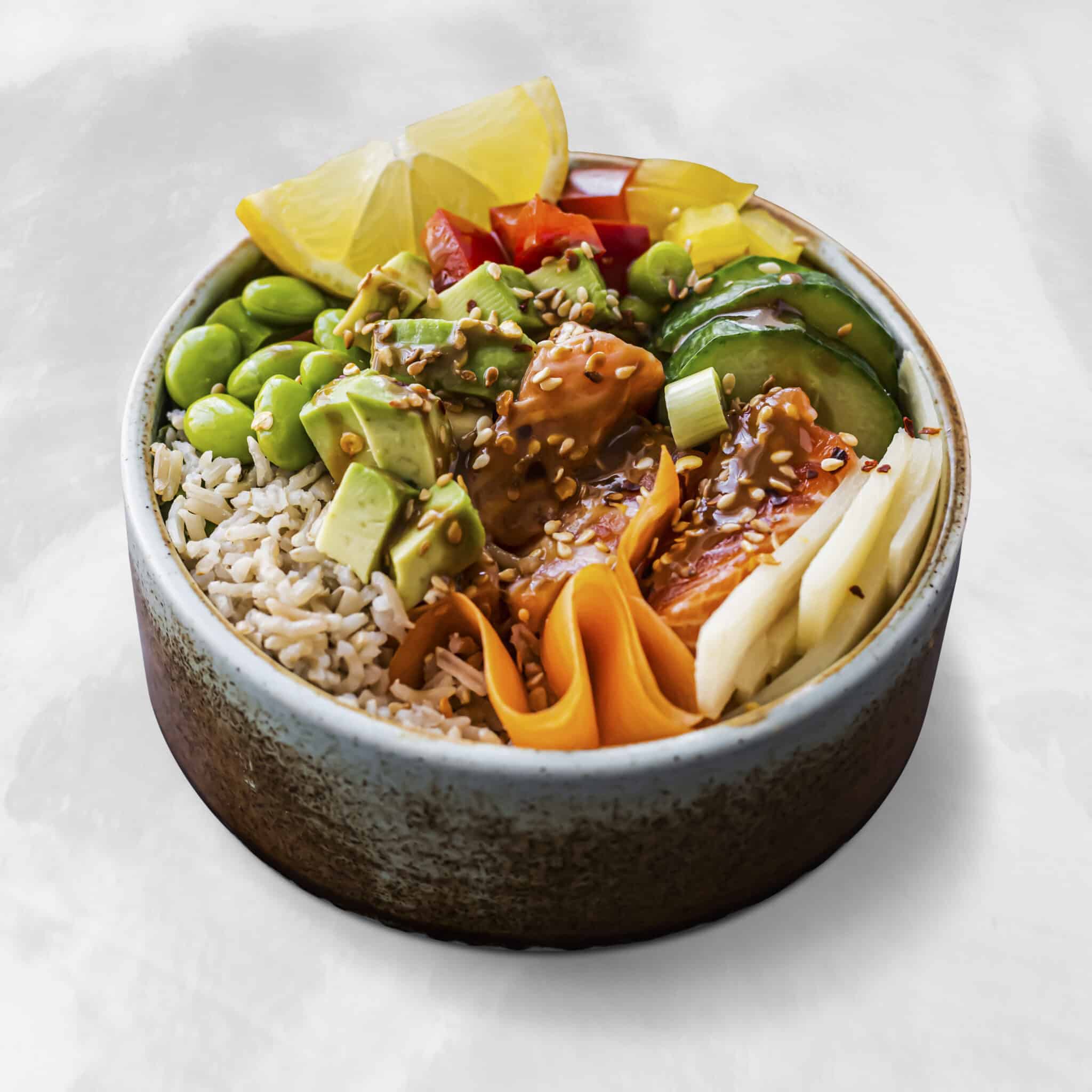 Close-up of a colorful poke-style bowl with rice, avocado chunks, edamame, pineapple, shredded carrot, cucumber, sesame seeds, and a savory sauce/condiment visible on top.