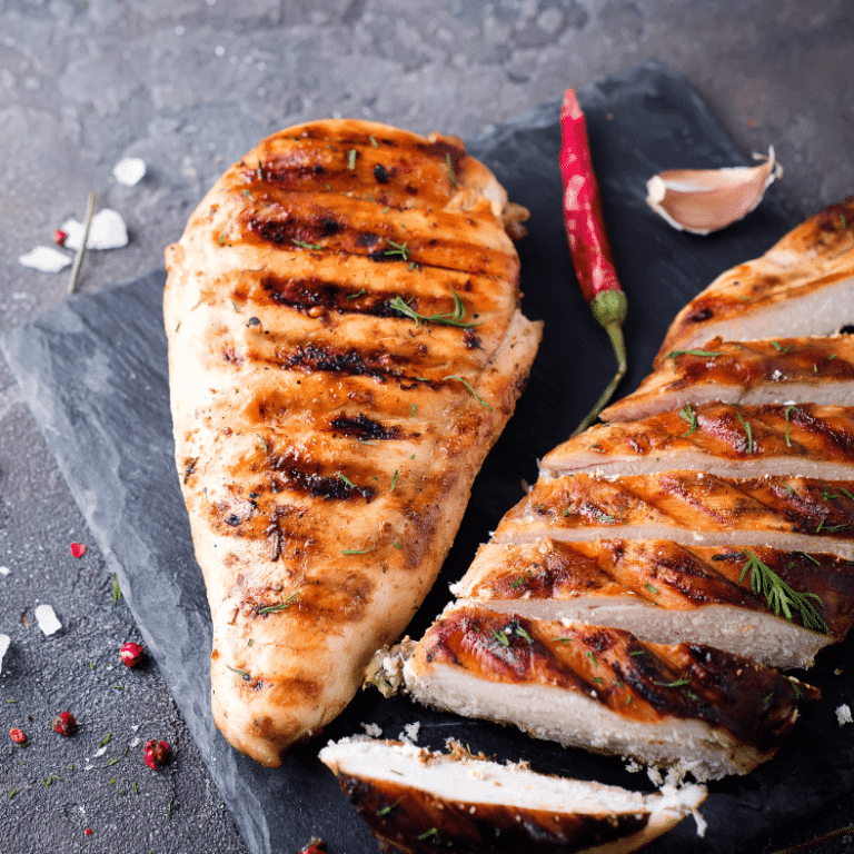 Grilled chicken breast and sliced chicken on a slate board, sprinkled with herbs and a chili pepper nearby.