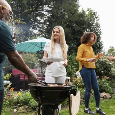 People enjoy a backyard barbecue: a man in a green shirt grills meat while a woman in a white star-printed top holds a plate among other guests chatting nearby by a grill and blue umbrella in a garden.
