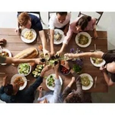 Top-down view of a group of people seated around a wooden table, sharing a meal with bowls and plates of salad and pasta in the center.