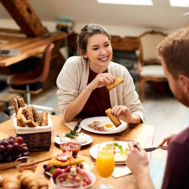 Happy couple eating breakfast and talking at dining table in the morning. Focus is on woman.