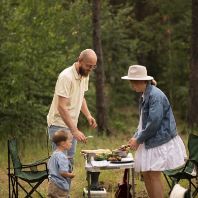 A family picnic in the woods: man, woman, and young boy cooking and serving food at a small outdoor table surrounded by camping chairs.