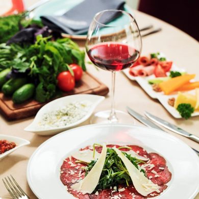 Plate of beef carpaccio topped with arugula and parmesan shavings on a dinner table, with a glass of red wine in the foreground.