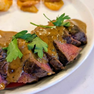 Sliced steak with brown sauce and fresh parsley garnish on a plate, with roasted potatoes in the background