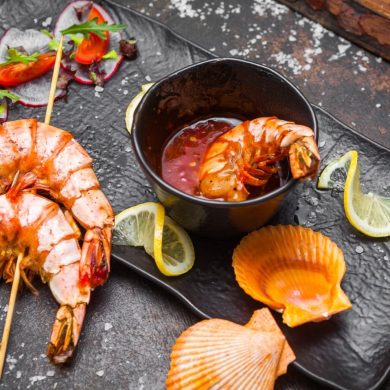 Platter of grilled shrimp with a dipping sauce in a black cup, lemon wedges, and seashells on a dark slate plate with garnish slices of radish and greens.