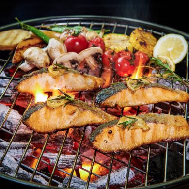 Salmon fillets grilling on a barbecue with lemon wedges, herbs, and vegetables over hot coals.