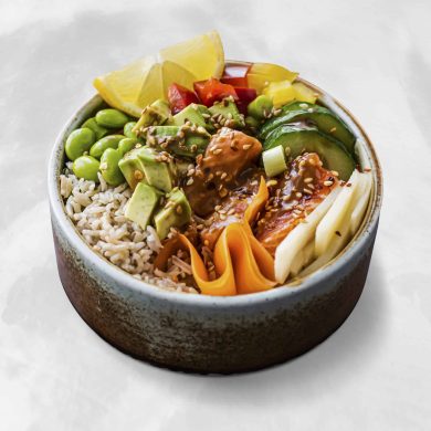 Close-up of a colorful poke-style bowl with rice, avocado chunks, edamame, pineapple, shredded carrot, cucumber, sesame seeds, and a savory sauce/condiment visible on top.