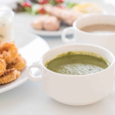 Plate of crispy fried chicken with a bowl of green soup and a white mug of coffee on a bright table surface.