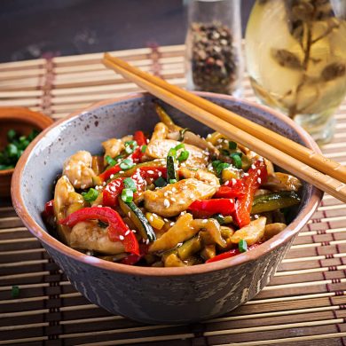 Bowl of chicken stir-fry with red peppers and greens, sesame seeds, and chopped scallions; chopsticks rest on the bowl's edge.